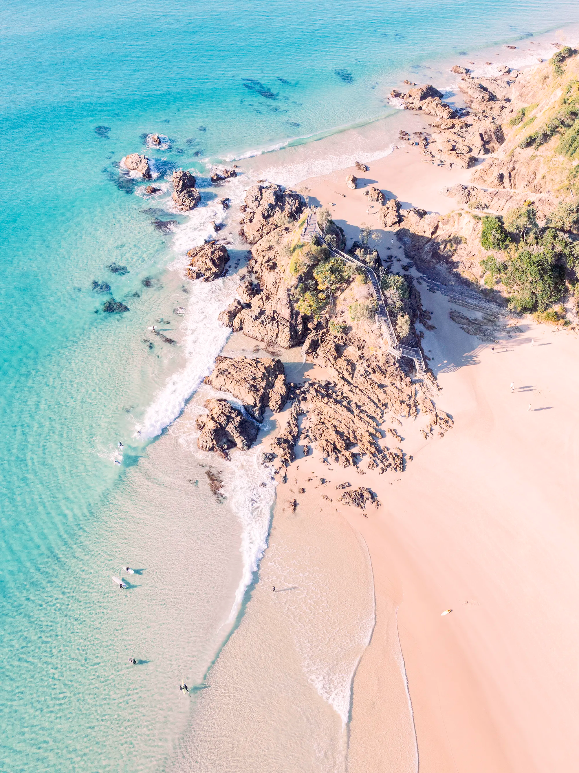 Aerial view of The Pass, Byron Bay showing rocky point with stairs, sand beach and shallow waves along a curved shoreline